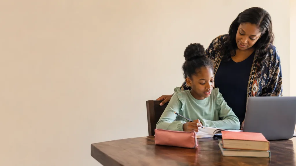A girl writing in her exercise book at a table while a woman stands beside her, looking on